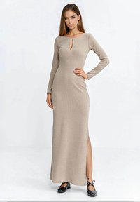 Beżowa sukienka z długim rękawem, z dekoltem w kształcie łezki, prążkowaną teksturą, bocznym rozcięciem i dopasowanym krojem. Nosi się ją z czarnymi płaskimi butami.