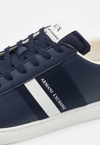 Sneakers blu navy con parte superiore in pelle liscia, dettagli in tessuto testurizzato, striscia bianca e marchio "Armani Exchange" di lato.