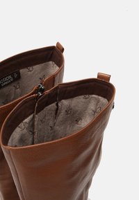 Bottes en cuir marron montantes avec doublure en tissu doux, dotées de détails de couture internes et d'une légère texture dans le matériau.