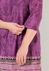 Lila Kurzarm-Tunika mit strukturiertem Muster, die am Saum dekorative horizontale Bänder mit geometrischen Designs aufweist.