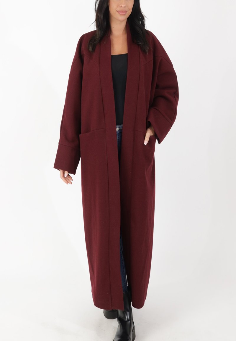 Abrigo largo granate hecho de tela gruesa con mangas anchas, apertura frontal y dos bolsillos delanteros. Se lleva sobre una camiseta negra y vaqueros azules.