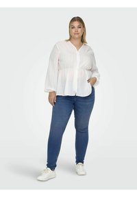 Bijela bluzica dugih rukava s gumbima sprijeda i suptilnim vertikalnim prugama, uparena s tamnoplaviim uskim trapericama i bijelim tenisicama.