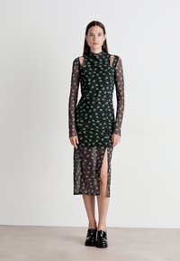 Long-sleeve, dark green dress with floral patterns, sheer overlay, high neckline, and side slit; worn with black footwear.