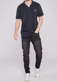 Granatowa koszulka polo z kołnierzykiem, krótkimi rękawami i małym logo na piersi, zestawiona z dopasowanymi czarnymi jeansami i białymi trampkami.