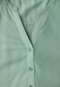 Camicia verde menta con scollo a V, patta con bottoni e maniche corte, realizzata in tessuto morbido con una texture liscia e un leggero lucido.