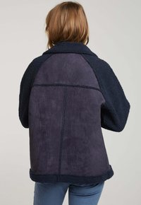 Veste bleu marine avec un haut en polaire texturée et un bas en suède lisse. Elle possède un col, des manches raglan et des détails cousus.