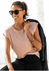 Light pink short-sleeve top with "VENICE BEACH" text, paired with black pants and layered necklaces. Leather jacket draped over shoulder.