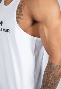 A fehér ujjatlan póló kiemeli az izmos felkar kontúrját, fekete "GORILLA WEAR" logóval rendelkezik. Az anyag simának tűnik, lágyan hullámzik.