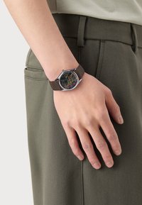 Brown leather strap watch with a round silver case, featuring a black face decorated with colorful abstract patterns.