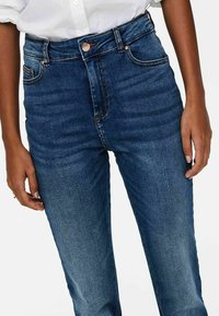 Gros plan sur une personne portant un jean bleu foncé taille haute et une chemise blanche boutonnée, les mains relâchées le long du corps.