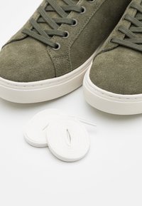 Olivgrön mockaskor med vita gummisålar och matchande snören. Har runda snören och en slät, mjuk yta.