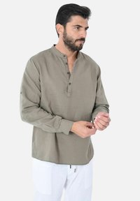 Camicia in lino verde oliva leggera con colletto rialzato, pattina con bottoni e maniche lunghe. Vestibilità su misura e orlo leggermente curvo.