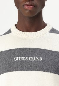 Szürke és krémszínű csíkos pulóver bordás kerek nyakkal. Az elején fehér hímzett felirat található: "GUESS JEANS". Texturált kötött anyag.