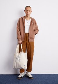 Hnědá mikina na zip s bílými akcenty, bílý tílko, hnědé kalhoty, šedé a tyrkysové tenisky a béžová taška přes rameno s modrým logem NY.