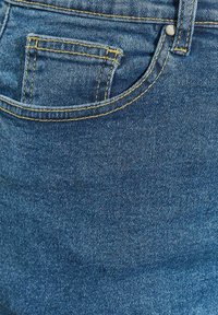 Tessuto in denim blu con una tasca posteriore visibile caratterizzata da dettagli di cucitura gialla. La texture appare leggermente usurata con una finitura liscia.