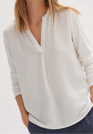 Vrouw draagt een witte blouse met lange mouwen en een V-hals met split, mouwen licht opgestroopt, handen deels in de zakken, gecombineerd met een donkere broek.
