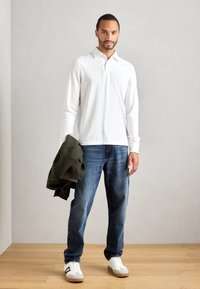 Mann mit einem weißen Langarmshirt, blauen Jeans und weißen Turnschuhen, der eine dunkelgrüne Jacke in einem minimalistischen Raum mit hellen Holzböden hält.