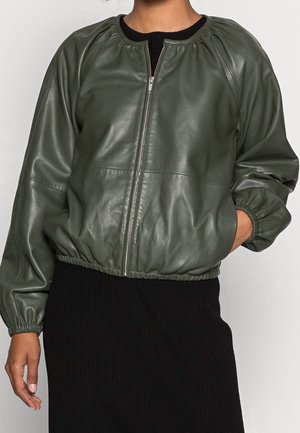 Femme portant une veste en cuir zippée vert foncé avec poignets et ourlet élastiques, sur une jupe noire côtelée, posant avec une main dans la poche.