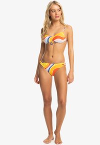 Kobieta modelka stojąca boso, ubrana w kolorowy paskowany bikini w pomarańczowe, żółte, niebieskie i białe spirale na białym tle.
