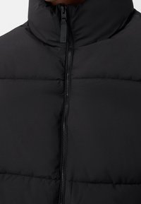 Svart puffer-väst med hög krage, quiltad textur och fullständig frontdragkedja. Materialet verkar mjukt och isolerat med minimala detaljer.
