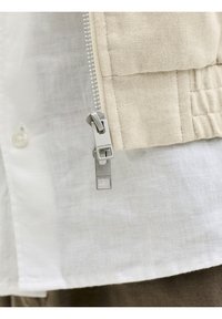 Gros plan d'une fermeture éclair argentée sur tissu beige au-dessus d'une chemise en lin blanc montrant la texture et les détails de couture.