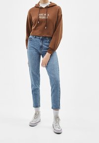 Persona que lleva una sudadera marrón con el texto "CONVÉRTE EN UNA PRIORIDAD", jeans de tiro alto azules, calcetines blancos y zapatillas blancas de caña alta.