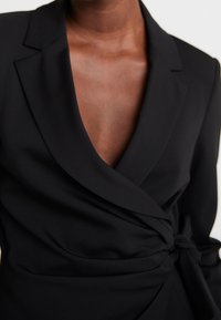 Svart omslag blazer med djup v-hals, strukturerade slag och knäppning fram. Slät tyg med minimal detaljer och skräddarsydd passform.