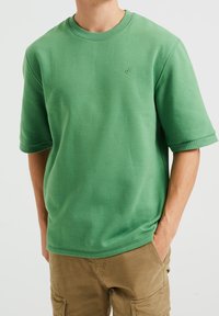 Zelená mikina s kulatým výstřihem vyrobená z bavlněné směsi, s krátkými rukávy a malým vyšívaným logem na levé části hrudi. Hladká textura.