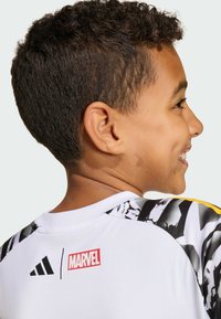 Hvid atletisk skjorte med sorte og gule detaljer, der har et tekstureret grafisk design på skuldrene og logoer fra Adidas og Marvel.