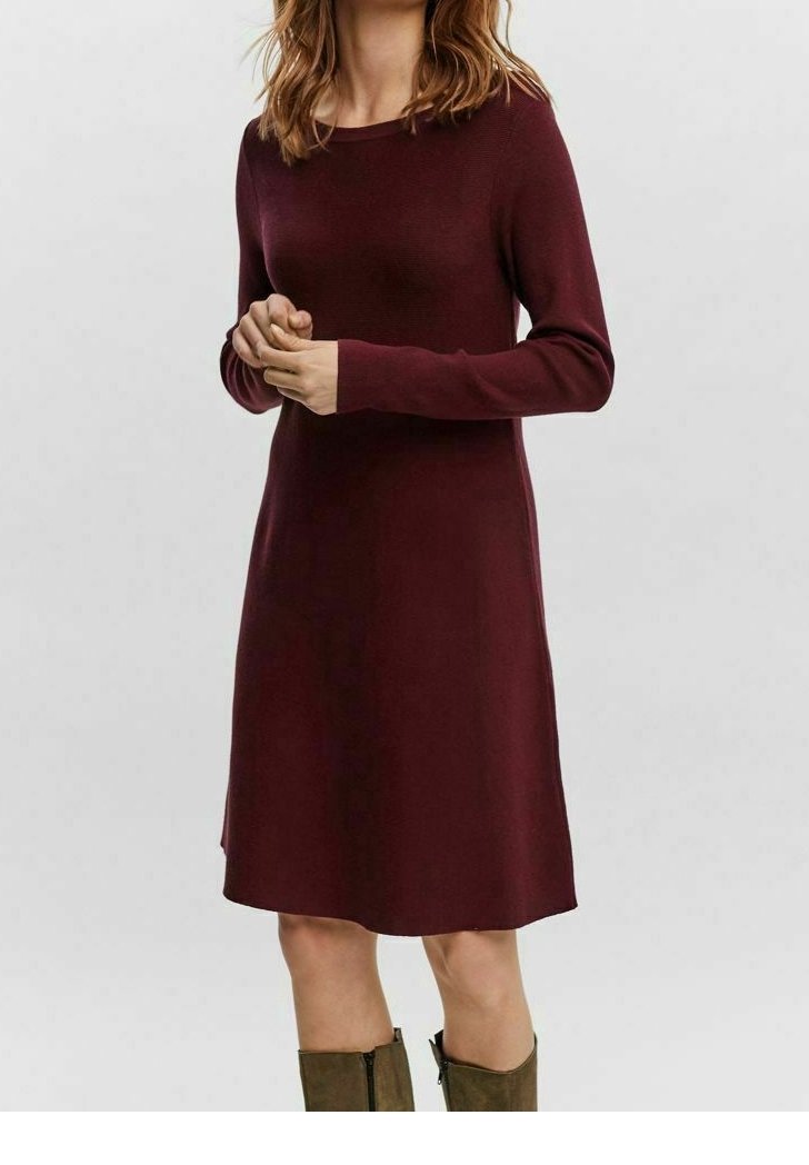 Femme portant une robe bordeaux à manches longues, longueur genou, et des bottes vert olive, debout, les mains légèrement jointes.