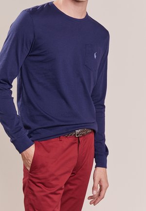 Homme portant une chemise bleu marine à manches longues avec une poche poitrine et un pantalon rouge avec une ceinture tressée marron, main dans la poche.