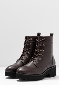 Bottes chevilles en cuir marron avec lacets, bout arrondi, semelle épaisse en caoutchouc noir et œillets métalliques. Équipées d'une languette à l'arrière du talon.