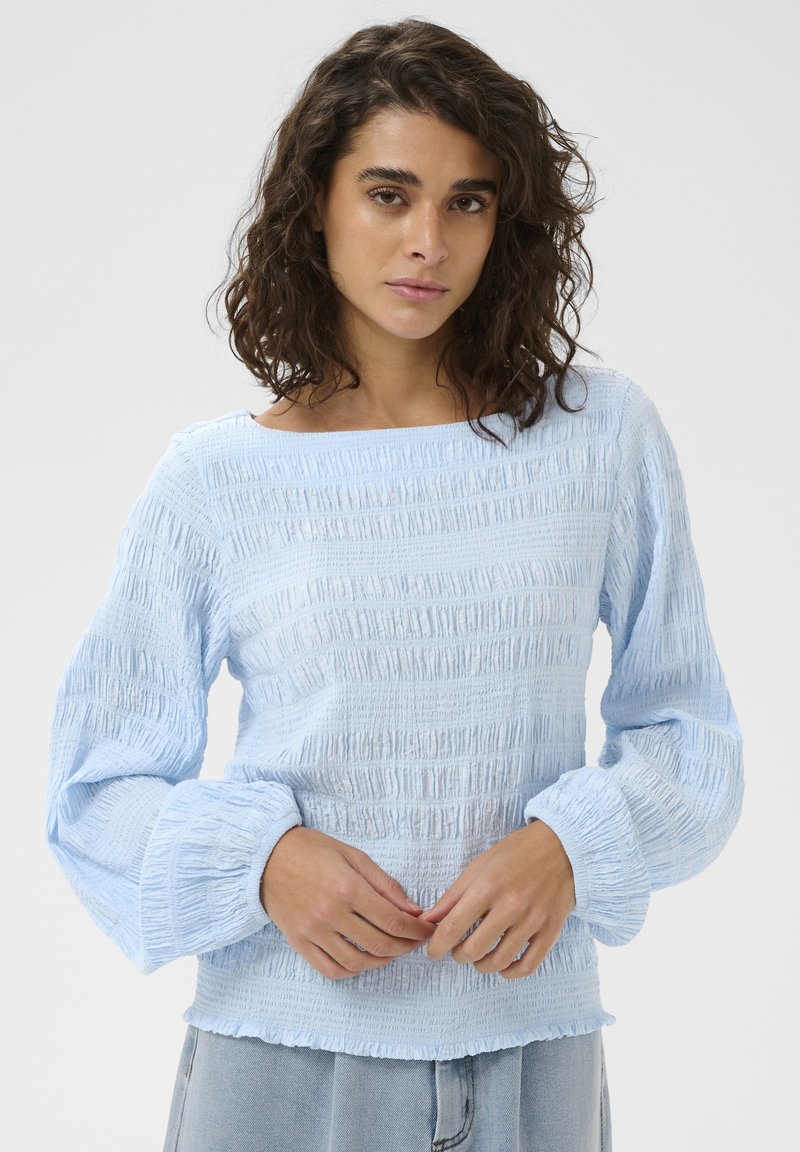 Jeune femme aux cheveux bruns bouclés portant un chemisier bleu clair texturé à manches longues et un jean en denim clair, debout devant un fond uni.