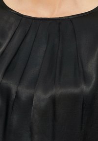 Schwarze Bluse aus einem glatten, glänzenden Material mit Falten am Ausschnitt und einem einfachen, runden Kragen.