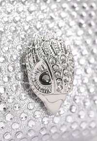Zilveren handtas met een getextureerde, glanzende oppervlakte. Voorzien van een metalen, ornamentale sluiting die lijkt op een oog, versierd met strass-stenen.