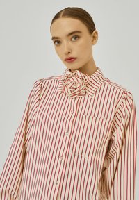 Gestreept, knoopshirt in roomkleur met rode verticale strepen. Heeft een grote rozenvormige accent bij de kraag en een borstzak.