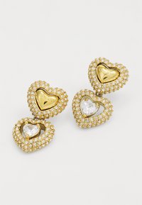 Boucles d'oreilles en or en forme de cœur ornées de strass transparents, avec une découpe centrale en forme de cœur et un précieux gemme, ainsi qu'un gravure de marque.
