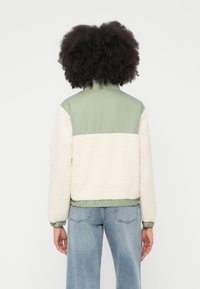 Kreemja fliisist jakk helerohelise nailonist yoke'i ja äärisega. Omadusteks on imitatsioonkrae ja elastsed varrukaotsad, ristkülikukujuline vorm ning soonikuga alläär.