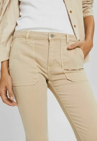 Beżowe dżinsy bawełniane o dopasowanym kroju, pięć kieszeni i widoczne detale ściegów. Gładka struktura z klasycznym pasem i zapięciem na guzik.