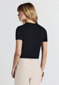 Donna con capelli biondi che indossa un top nero a maniche corte e pantaloni beige a vita alta, girata di spalle contro uno sfondo chiaro e uniforme.