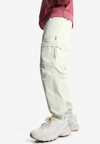 Oseba v off-white cargo hlačah z elastičnimi manšetami, roza sweatshirtu, belih crew nogavicah in belih chunky športnih čevljih, stoji bočno.