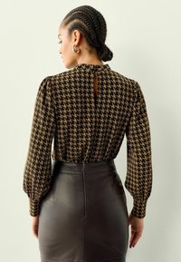 Bluza s hounds-tooth uzorkom u crnoj i zlatnoj boji, s puf rukavima i detaljem u obliku ključa na leđima, u kombinaciji s uskom kožnom suknjom.