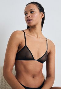 Zwarte doorschijnende driehoekige bralette met dunne bandjes, met een klein detail van zilveren hardware in het midden. Gladde textuur en minimalistisch ontwerp.