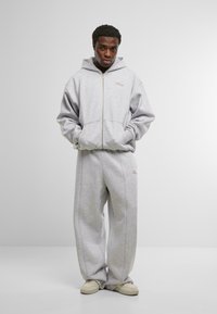 Felpa grigia con cappuccio e zip frontale, abbinata a pantaloni in felpa a gamba larga. Entrambi presentano un logo discreto. Texture morbida.