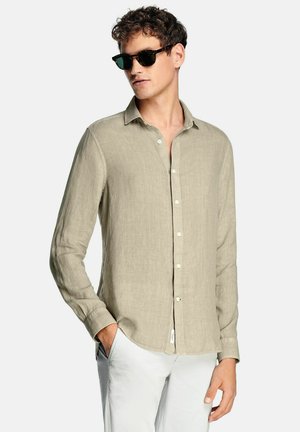 Man draagt een beige lange mouwen overhemd met knopen, witte broek en zwarte zonnebril, staat met één hand in de zak.