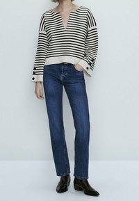 Personne portant un pull à col en V rayé noir et blanc, un jean bleu à jambes droites et des bottines noires, debout avec une main dans la poche.