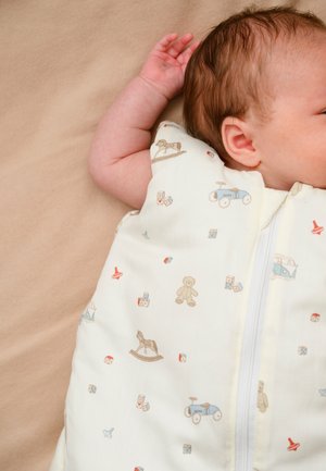 Bebeluș adormit, purtând un sac de dormit alb cu imprimeuri mici de căluți balansoar, ursuleți de pluș, mașinuțe de jucărie și cuburi, pe o suprafață bej.