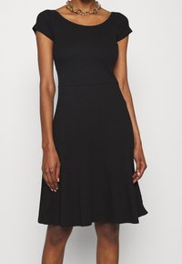 Svart klänning med en åtsittande bodice och utsvängd kjol, med korta ärmar som är något avkapade och en mjuk, elastisk tygstruktur.