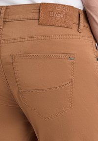 Bruine stretch stof broek met een gladde textuur, voorzien van een leren label met het "Brax" logo en een achterzak met een klein logo label.