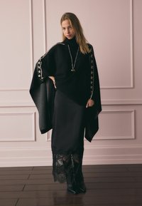 Fekete kötött poncsó csillagmintás díszítéssel, hosszú fekete szoknyával, amely csipke szegéllyel és fekete, magas sarkú csizmával van párosítva. Fa padló háttér.